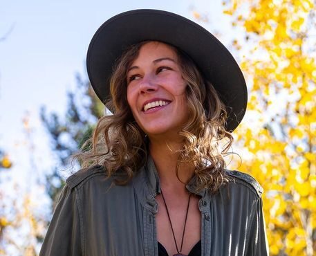 A joyful woman with curly hair wearing a black hat and olive green shirt smiles under a canopy of golden autumn leaves.