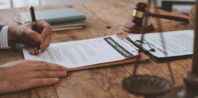 Two people are seen signing a contract agreement on a clipboard on a wooden table. Nearby are a gavel and a pair of scales, suggesting a legal or professional setting. One person holds a pen while another's hands rest on the document.