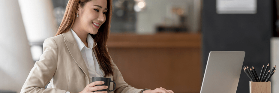 A person with long hair wearing a light-colored blazer and white shirt is sitting at a desk, smiling while working on a laptop. They are holding a mug in one hand. The background shows a blurred office space with a container of pencils to one side.