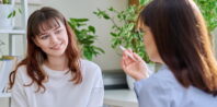 Two women are sitting and talking in a bright room filled with plants. One woman with brown hair and bangs is smiling, wearing a light-colored sweater. The other, her associate with darker hair, blurred and wearing glasses and a striped shirt, gestures animatedly with her hand.