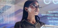A woman with glasses looks at digital screens displaying code, reflecting a blend of focus and technology in a modern setting.