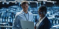 Two professionals, a man and a woman, smiling and discussing over a laptop in a server room with rows of computer racks in the background.