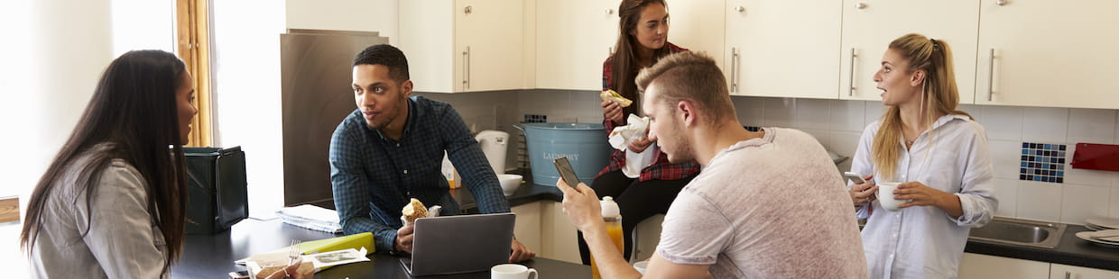A group of five college students are gathered in a modern kitchen. Two are sitting on the counter, two are standing, and one is sitting at a table with a laptop and smartphone. They appear to be conversing and eating. The bright kitchen is equipped with white cabinets, reflecting their discussion on food access.