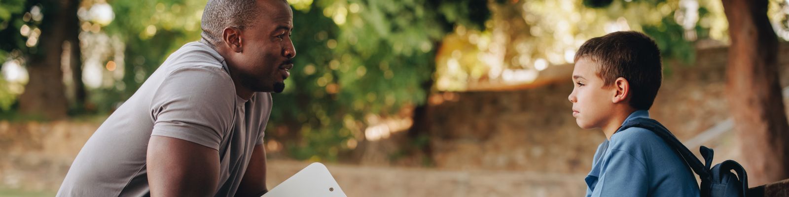 A man kneels while talking to a young boy wearing a backpack outdoors. The man, possibly discussing social work education, is holding a white clipboard, and both appear engaged in conversation. The background is filled with greenery and sunlight.
