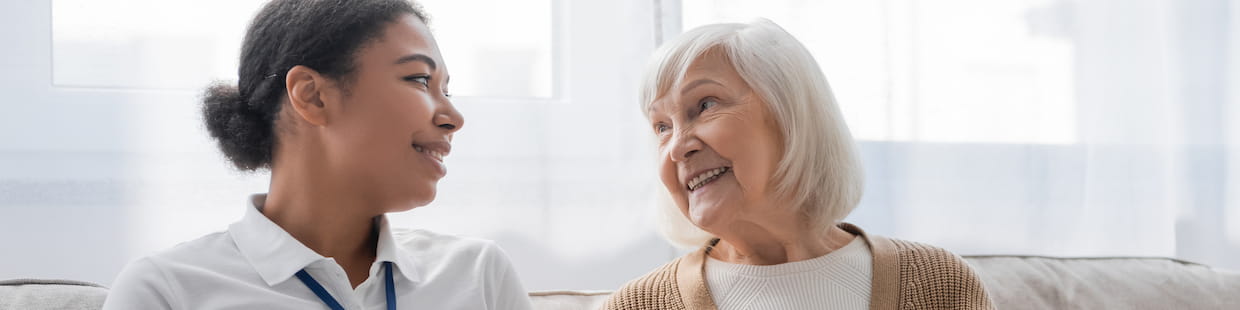 An elderly woman with white hair and a beige sweater smiles warmly while looking at a younger woman with dark hair tied back, who wears a white shirt and a name tag displaying her credentials. They sit on a sofa in a bright, sunlit room, enjoying each other's company.