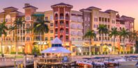 A scenic waterfront view of a vibrant, multi-story building complex at sunset. Palm trees line the front of the buildings, which feature a mix of colorful facades. In the foreground, boats are docked at a marina, enhancing the tranquil atmosphere.