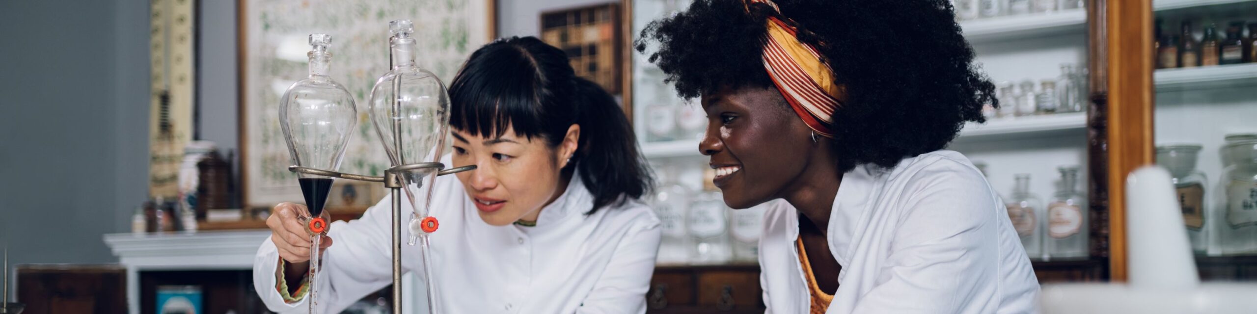 Two scientists, wearing white lab coats, work in a laboratory. The woman on the left is adjusting an important glass instrument, while her associate watches, smiling. Behind them, shelves are filled with various glass containers and lab equipment.