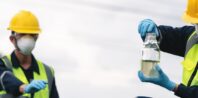 Two workers in high-visibility vests, hard hats, and protective masks and gloves work outdoors. One associate holds a sample jar with water, inspecting it closely, while the other observes. The background includes trees and a blurred sky, suggesting an open environment.