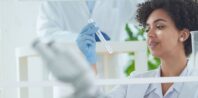 A lab technician with curly hair examines a test tube while wearing blue gloves and a white lab coat. An associate in the foreground, slightly out of focus, looks on. The background shows lab equipment and a blurred colleague in similar attire.