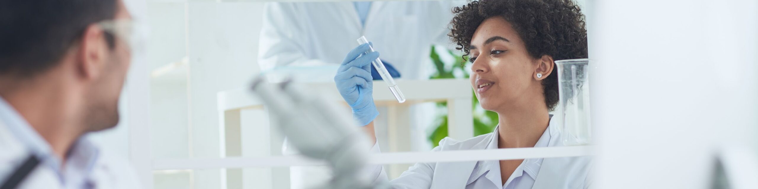 A lab technician with curly hair examines a test tube while wearing blue gloves and a white lab coat. An associate in the foreground, slightly out of focus, looks on. The background shows lab equipment and a blurred colleague in similar attire.