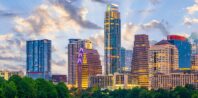 Panoramic view of a city skyline at sunset, featuring modern high-rise buildings and skyscrapers with a backdrop of a partly cloudy sky. The foreground includes green trees, creating a contrast with the urban landscape.