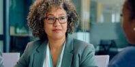Three people are engaged in a business meeting in a modern office. The woman in the middle, wearing glasses and sporting curly hair, is attentively listening. Another woman to her left is looking ahead, while the third person—whose back faces the camera—speaks about Graduate Certificates.