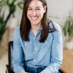 A woman with long brown hair, wearing a denim shirt and jeans, sits on a chair and smiles at the camera. There are plants in the background, creating a cozy and natural ambiance.