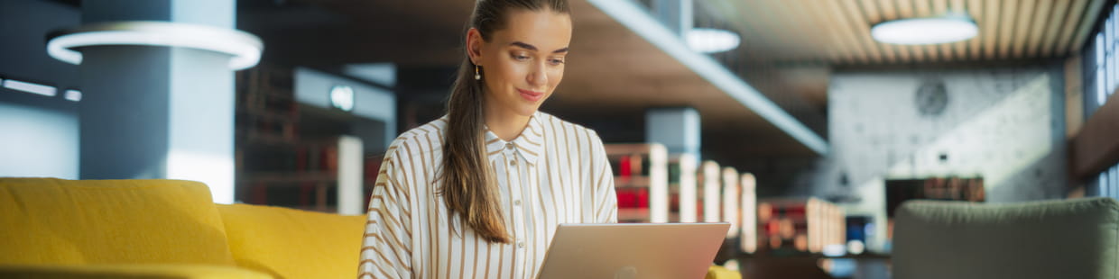 A woman, likely a recent bachelor's graduate, sits on a yellow sofa in a modern, well-lit room with wooden elements, using a laptop. She is smiling and appears focused on her work. The background features sleek architectural design with circular lights and a blurred shelf or cabinet.