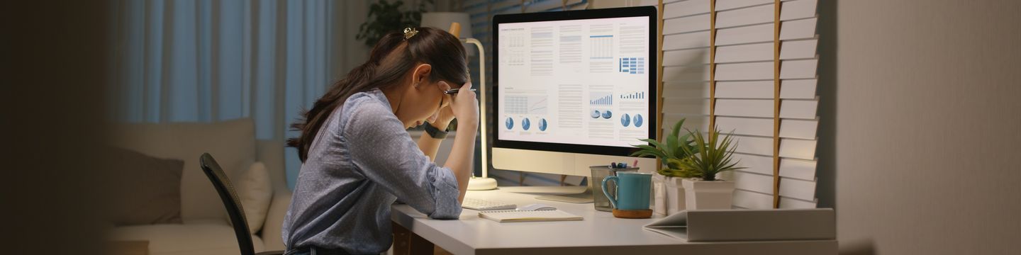 A person sits at a desk looking at a computer monitor displaying graphs and charts. They rest their forehead on their hand, appearing stressed or tired. The desk has a lamp, a potted plant, a coffee mug, and a notebook. The background is a dimly lit room.