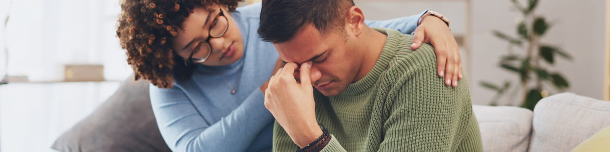 A person with curly hair and glasses comforts a grieving individual wearing a green sweater who is sitting on a couch, covering their face with one hand. The background features light-colored walls and a potted plant, echoing the sense of loss in the room.