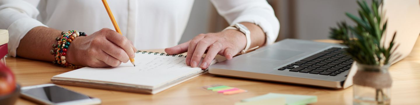 Close-up of a person writing in a notebook with a pencil. The person's hands rest on a wooden desk, alongside a laptop, sticky notes, a smartphone, and a small potted plant. The person is wearing a white shirt and multiple bracelets on one wrist.