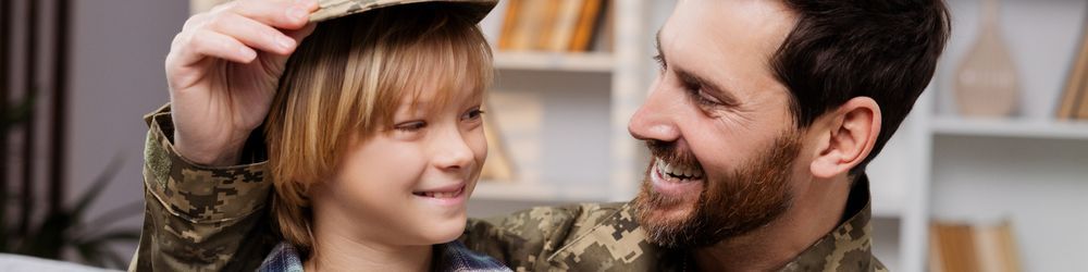 A person in military uniform smiles and adjusts the cap of a young child who is also wearing a similar uniform. They are indoors with bookshelves in the background and both appear happy.