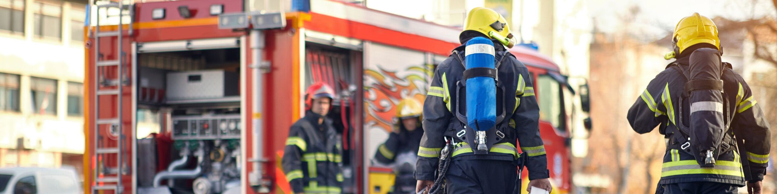 Three firefighters in full gear, including oxygen tanks, stand near a fire truck with its compartments open. The scene appears to be in an urban setting with buildings in the background. The firefighters, likely associates discussing a strategy or engaged in activity, convey a sense of readiness and teamwork.