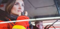 A woman in a red safety jacket is sitting in a vehicle, speaking into a radio with focused intensity as she looks ahead. An associate also wearing a safety jacket is visible in the background, their details blurred.