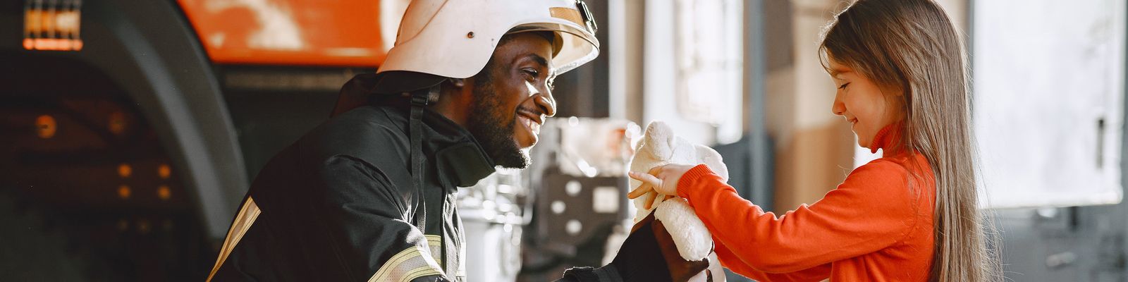 A firefighter in uniform kneels and smiles at a young girl who is handing him a white stuffed animal. Inside the fire station, with part of a fire truck visible in the background, they share a moment of connection. The girl, wearing a red shirt and with long brown hair, seems to associate safety with her hero.