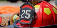 A red firefighter helmet with the number 25 on the front, surrounded by associated gear including gloves, belts, and reflective vests, lying on the ground. The background shows blurred firefighter equipment and boots.
