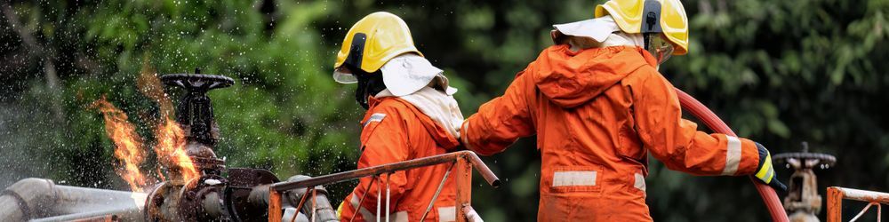 Two firefighters in orange protective gear and yellow helmets are handling a hose, aiming water at a small fire on industrial piping outdoors. Water droplets are visible in the air, while green foliage appears in the background.