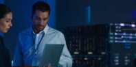 A woman and a man are standing in a dimly-lit control room, focused on a laptop screen. The background features large digital displays and data charts, indicating a high-tech environment. Other people are working in the background.