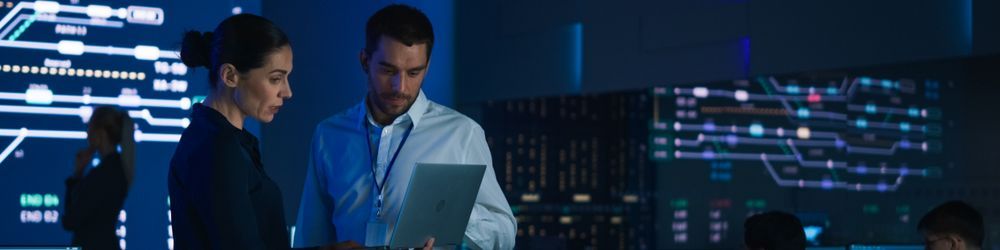 A woman and a man are standing in a dimly-lit control room, focused on a laptop screen. The background features large digital displays and data charts, indicating a high-tech environment. Other people are working in the background.