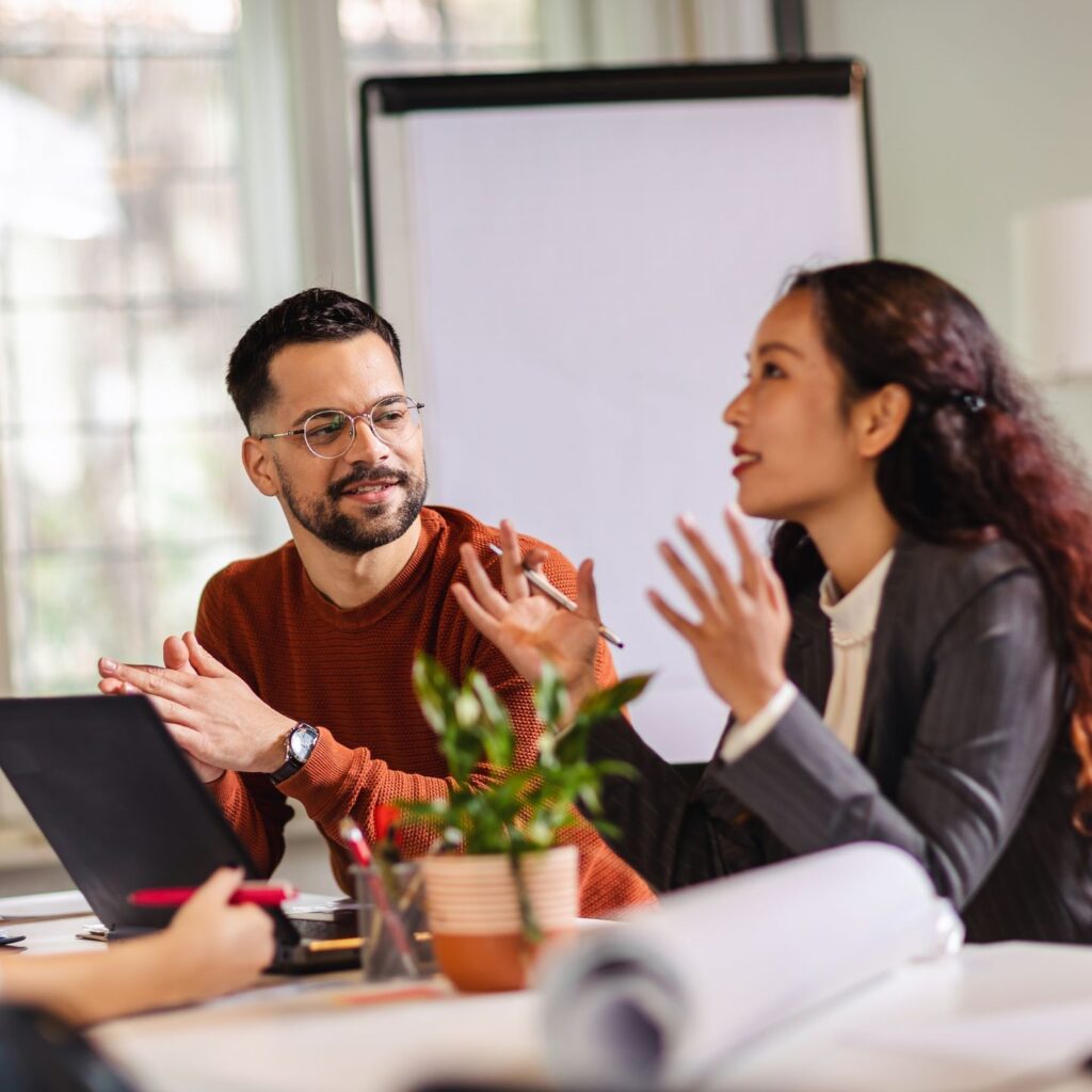 A woman in a gray blazer and a man in a brown sweater discuss editorial values at a table. The woman gestures with her hands while speaking. A laptop, potted plant, and roll of paper are on the table. A blank flip chart stands in the background.