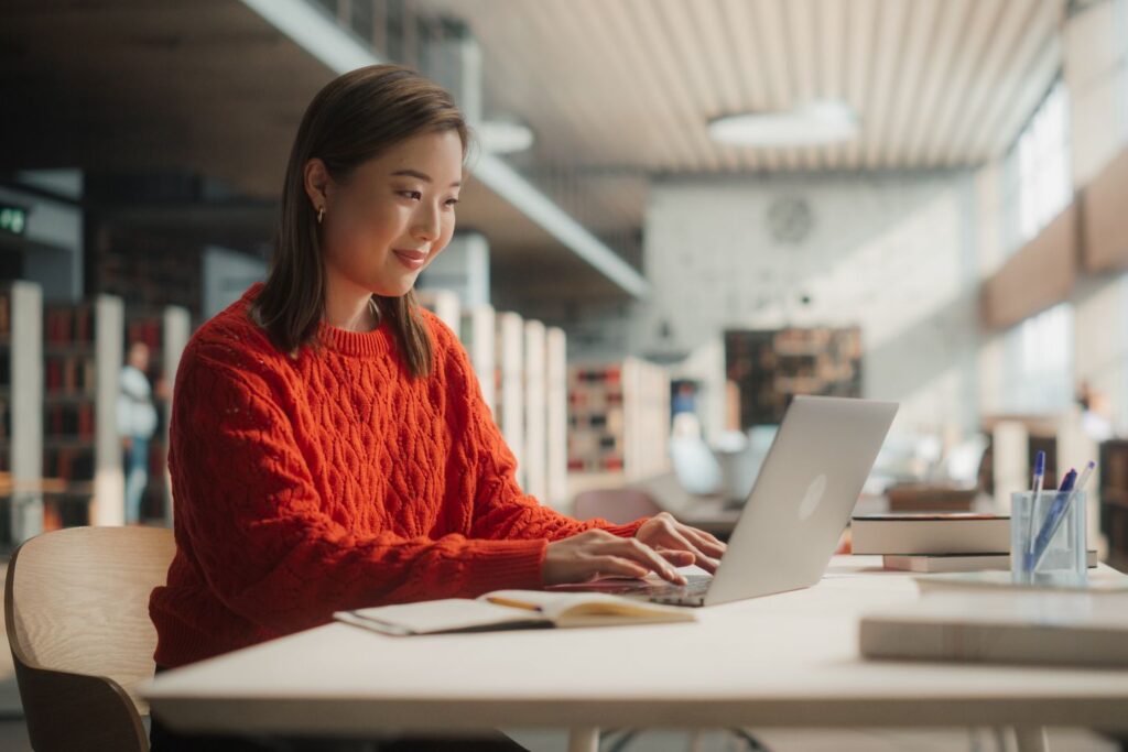 A person wearing a red sweater sits at a desk in a well-lit room with bookshelves. They are smiling and working on a laptop, surrounded by notebooks and a pen holder on the table. The background is slightly blurred, creating a warm, focused atmosphere that embodies strong editorial standards.