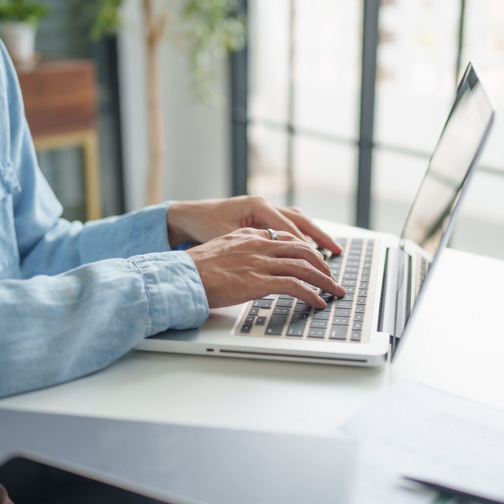 A person is typing on a laptop keyboard at a desk, embodying strong editorial values. They are wearing a light blue shirt, and the background shows a large window with blurred greenery outside. The workspace exudes a modern and bright atmosphere.