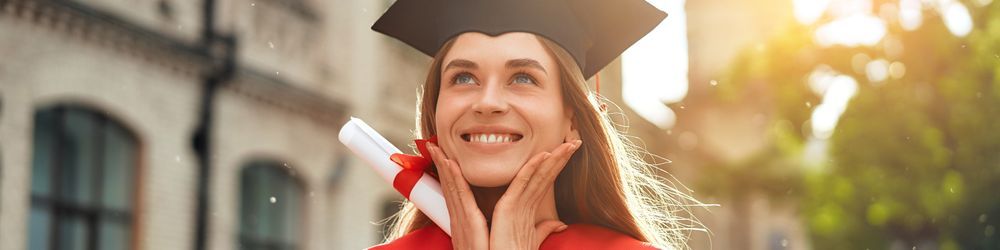 A young woman wearing a graduation cap and gown smiles brightly while holding a diploma tied with a red ribbon. Sunlight filters through trees in the background, creating a warm, celebratory atmosphere.