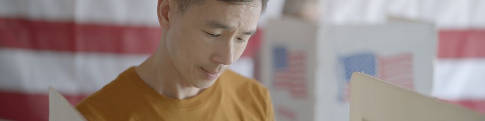 A person is standing in a voting booth, looking at a ballot. An American flag is visible in the background.