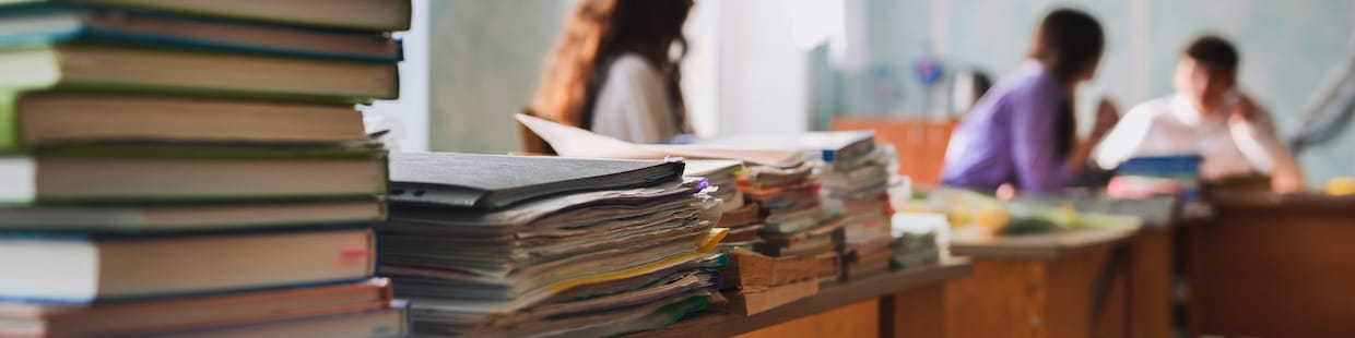 Stacks of books and documents clutter a table in the foreground. In the blurred background, three people are immersed in discussion about non-teaching career paths. The room exudes a casual, academic atmosphere.