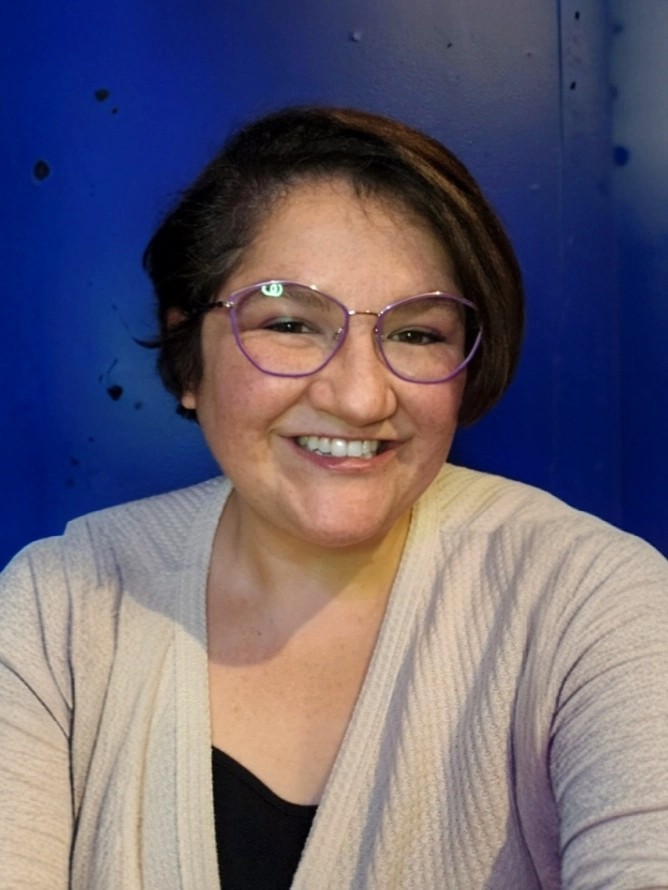 A person with short hair and glasses smiles at the camera. They are wearing a light beige cardigan over a black top. The background is a solid blue color with a few small black dots.