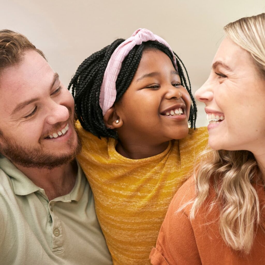 A smiling young girl with braided hair and a pink headband sits between two adults, all laughing and looking at each other, creating a joyful and affectionate moment.
