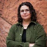 A person with curly hair and glasses wearing a green corduroy jacket over a black shirt stands with arms crossed against a textured reddish-brown stone wall.